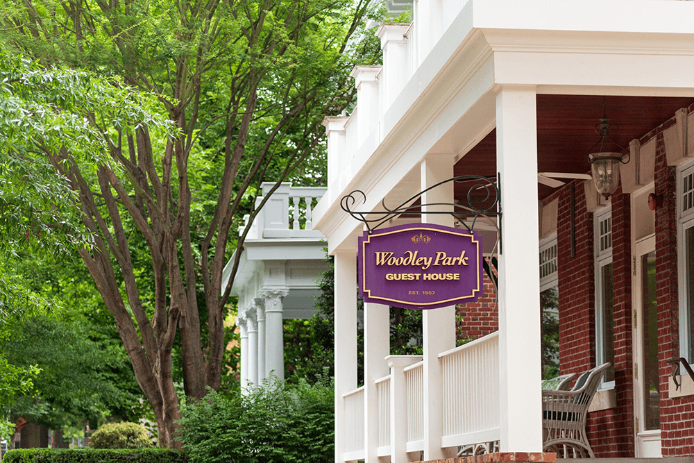 Sign for Woodley Park Guest House hangs on a white porch surrounded by green trees.