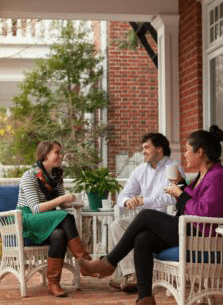 Three people enjoy a conversation on a porch with greenery in the background.