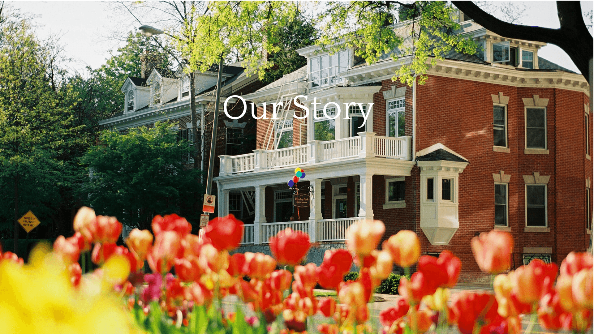 A brick building with white trim, surrounded by colorful tulips, features a sign reading "Our Story."