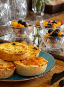 Four savory quiches on a blue plate, accompanied by bowls of mixed fruit.