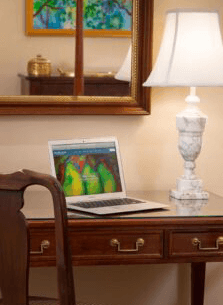 A laptop sits on a wooden desk beside a table lamp and a mirror reflecting a colorful painting.