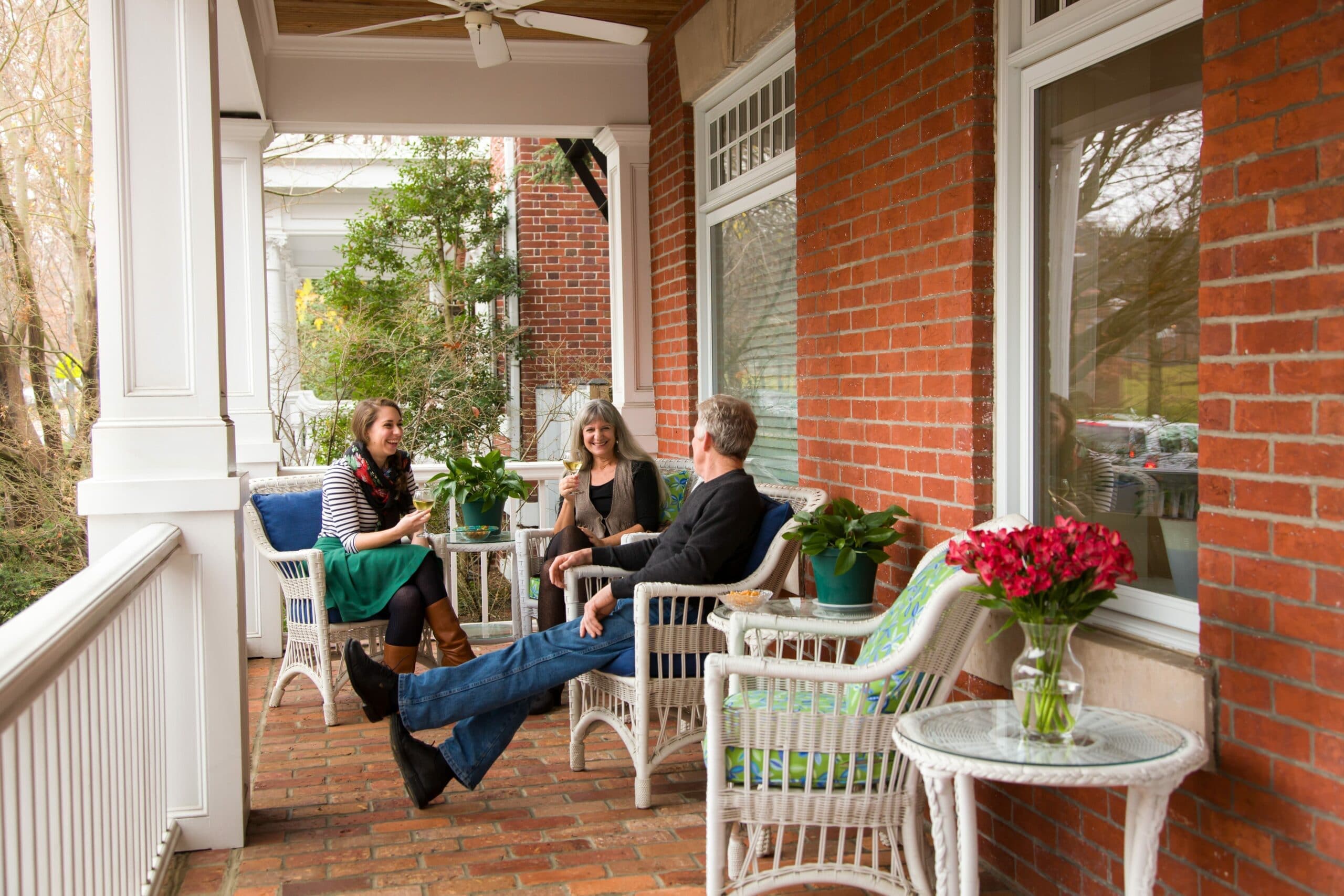 Three people enjoy drinks and conversation on a cozy porch surrounded by greenery.