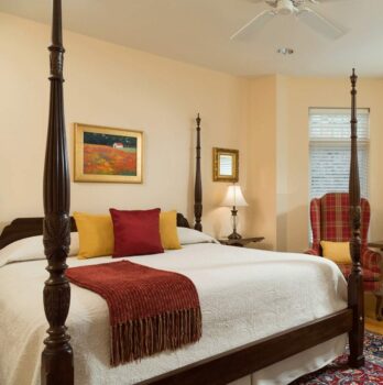 A room features large sunny windows, a four post king sized bed, and a comfortable seating area
